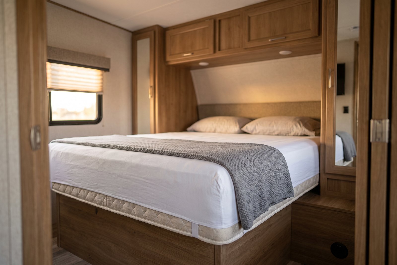 RV king mattress made up on a raised platform bed in a fifth-wheel front bedroom, with overhead cabinets and a slide-out wall